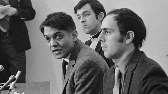 Eqbal Ahmad, (L) is shown as a spokesman for Father Philip Berrigan, where he told newsmen that Berrigan dismissed Ramsey Clark as his attorney in the Harrisburg 7 Trial that opened here. Berrigan had asked to make an opening statement, but was denied by the judge. When asked to defend himself for being denied, he explained that he considered himself without counsel. Left to right is Ahmad, Reverend Neil McLaughlin, and former priest Anthony Scoblick.